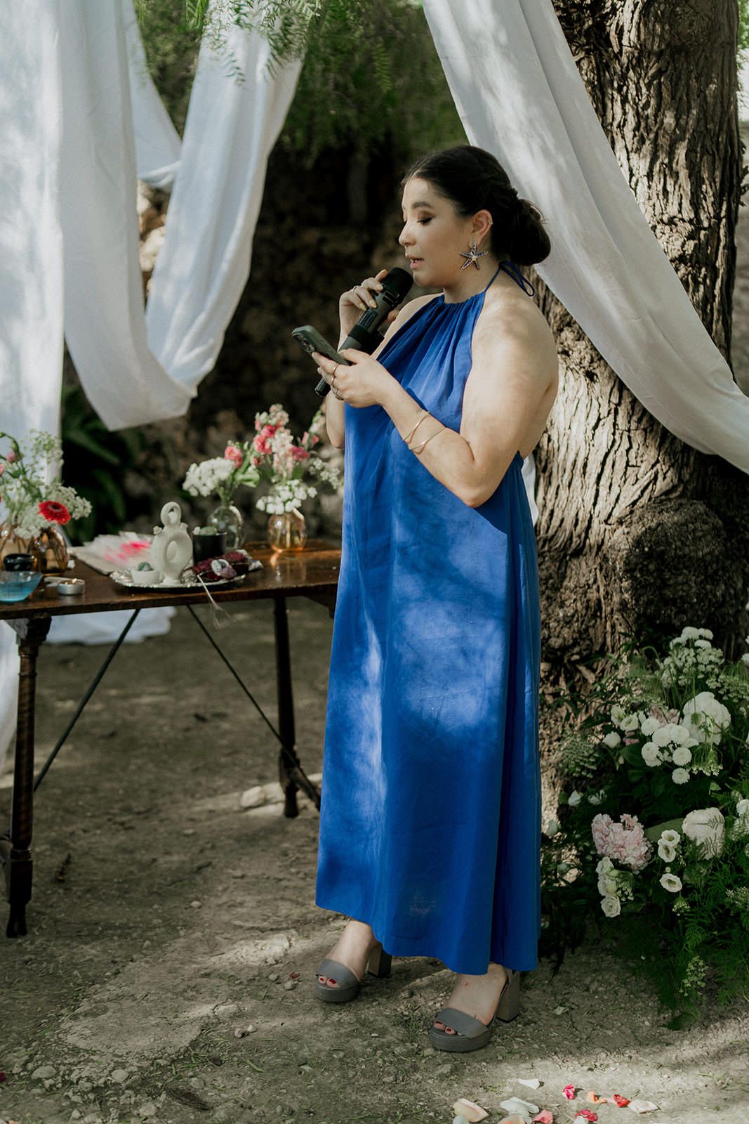 Person in vibrant blue dress speaking into microphone at outdoor garden wedding reception.