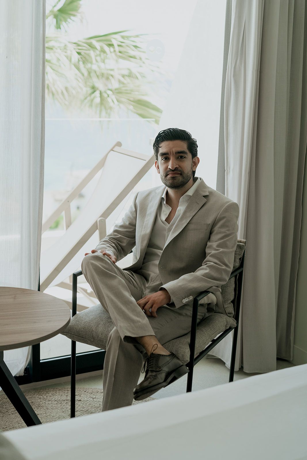 Man in light colored suit sitting in chair by window with palm tree view.