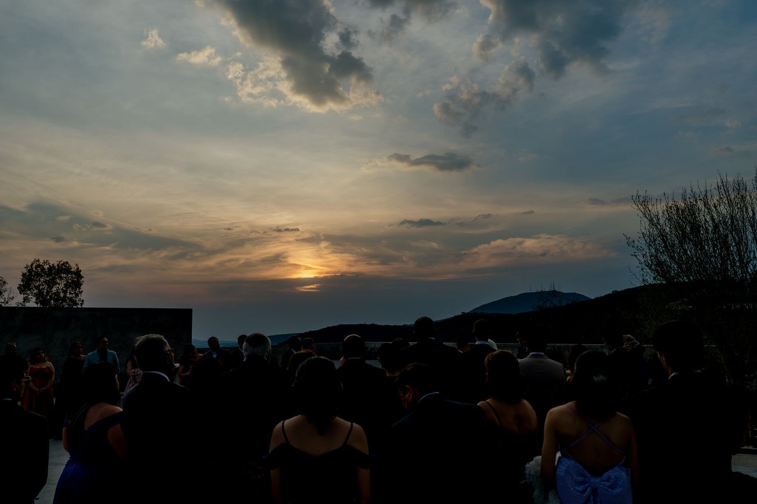 Dramatic sunset sky with golden clouds silhouettes wedding guests gathered at outdoor ceremony.