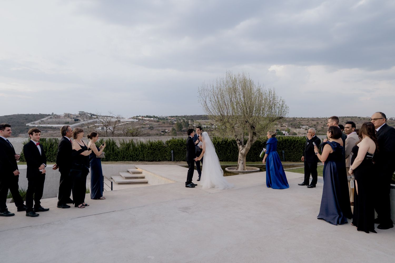 Outdoor wedding ceremony on a rooftop terrace with bride, groom, and guests under a cloudy sky.