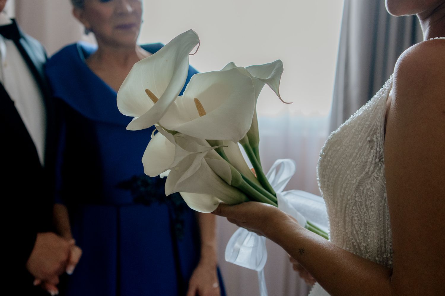 Bride holding elegant white calla lily bouquet at wedding ceremony with guests in blue dress background.