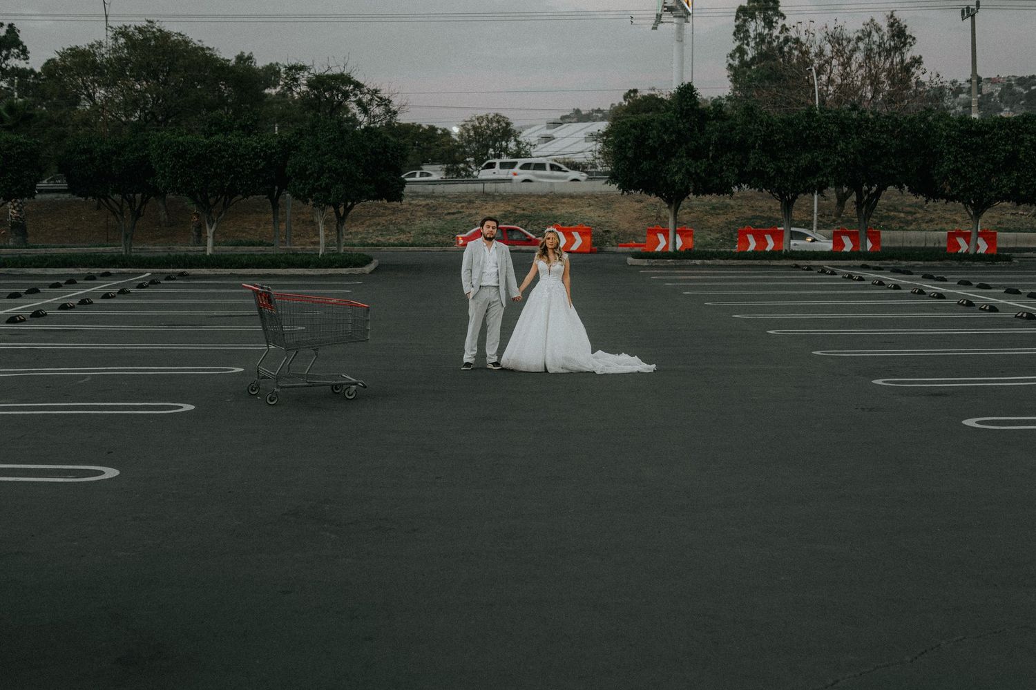 A couple poses with shopping carts in an empty parking lot with orange lights in the background.