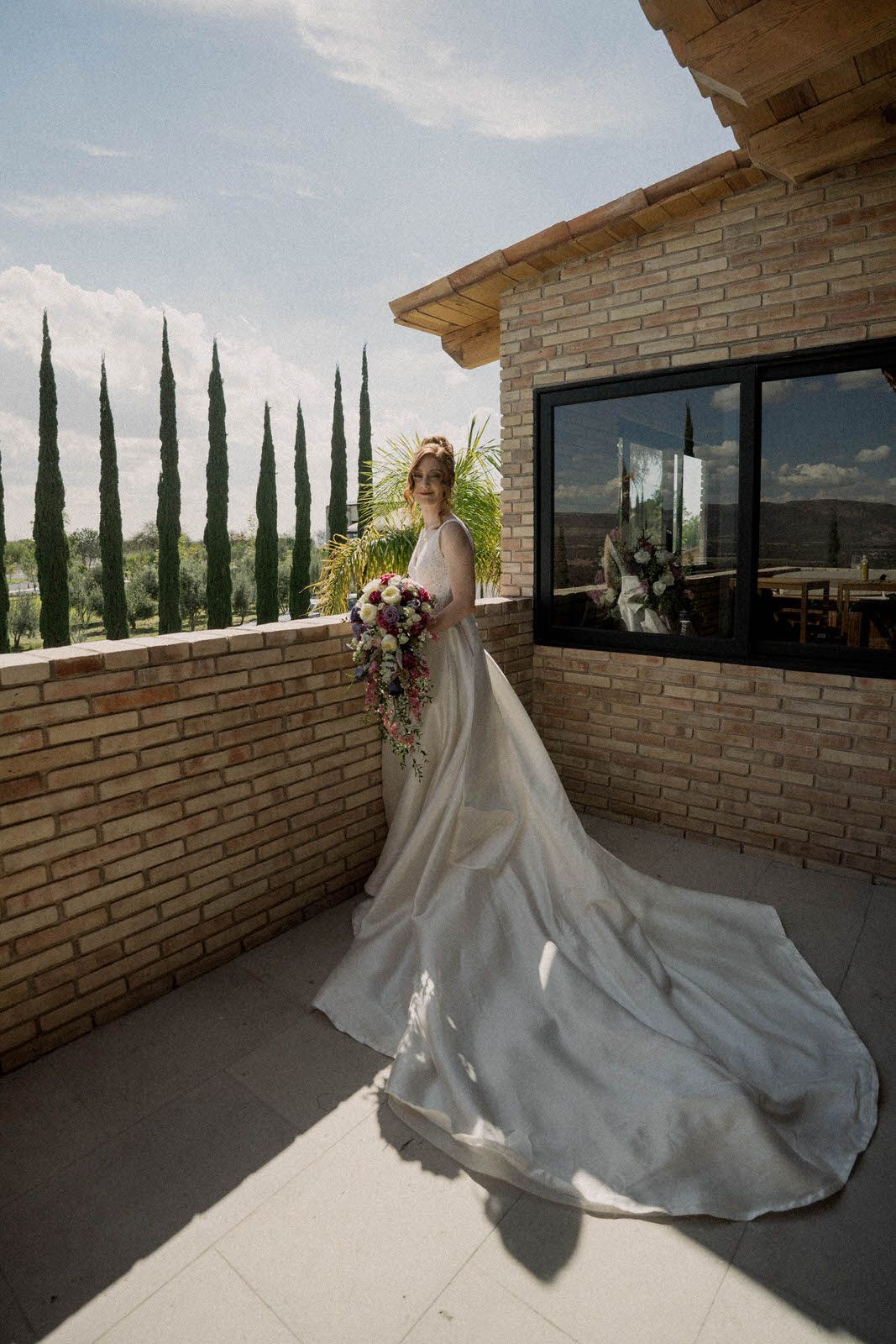 Elegant bridal gown with cathedral train photographed against brick and cypress backdrop.