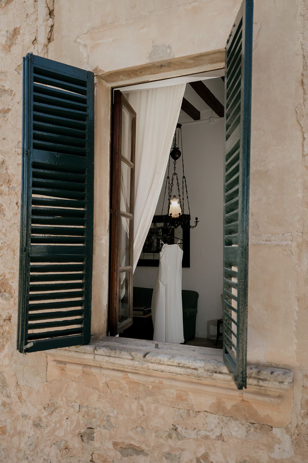 White dress visible through rustic window with green shutters and hanging chandelier.