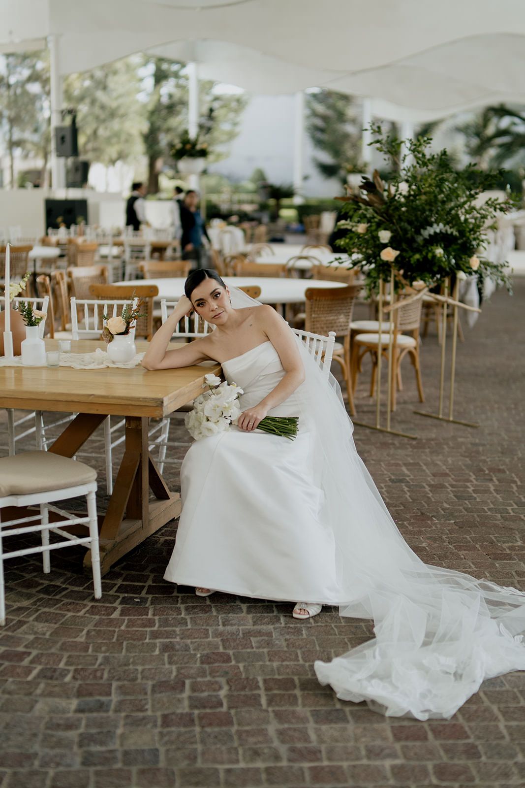 Elegant bride in strapless gown sits at wooden table holding white bouquet in decorated wedding venue.