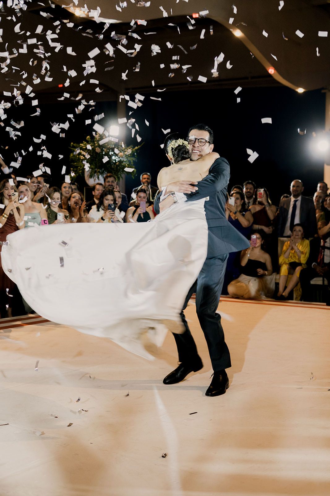 Groom lifts bride in stunning first dance moment as confetti fills the air with guests cheering in background.