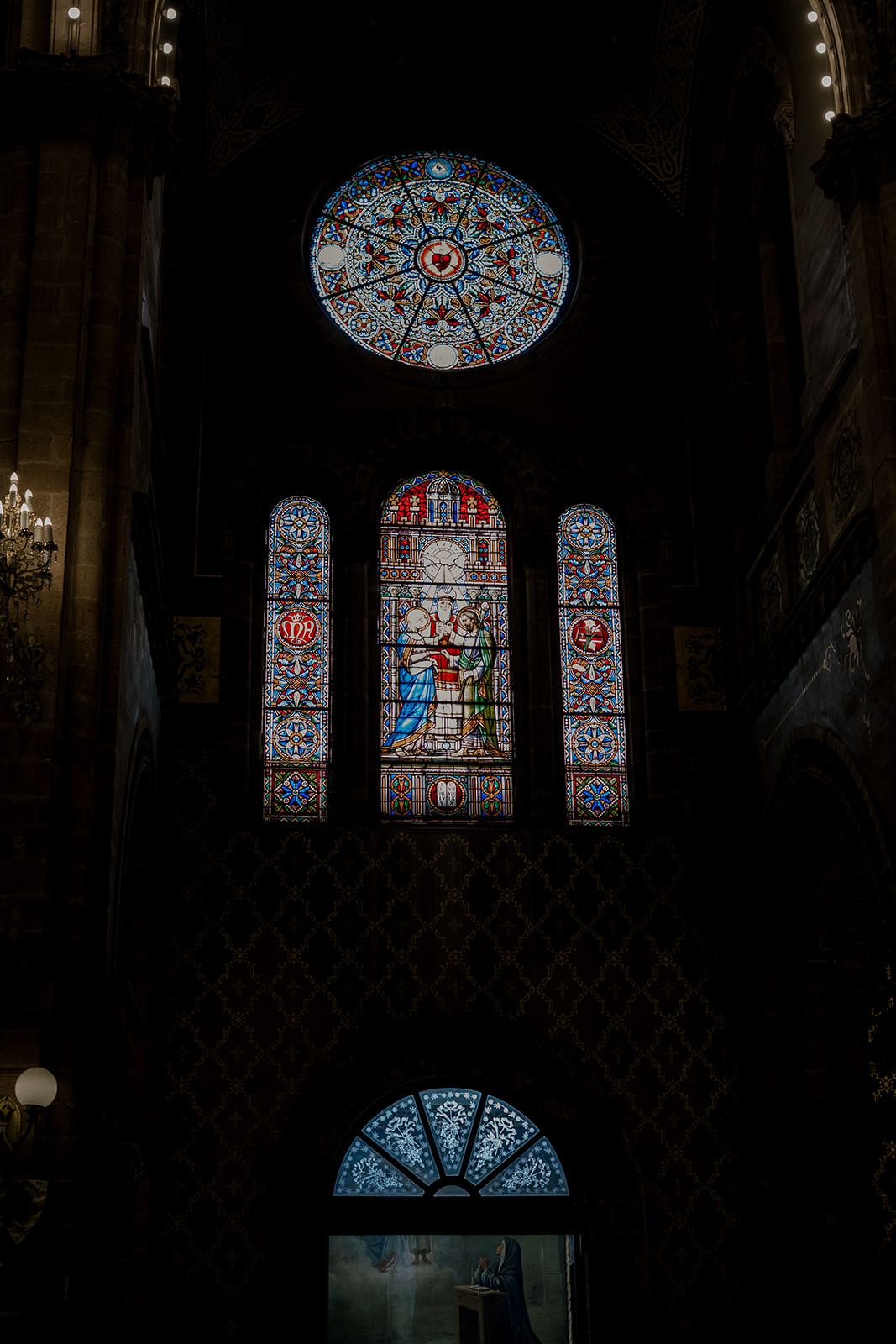 Stunning stained glass windows in a dark cathedral interior featuring colorful religious scenes and a circular rose window.