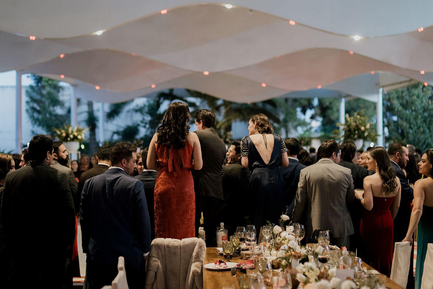 Wedding guests mingle under a draped tent with string lights and floral centerpieces at evening reception.