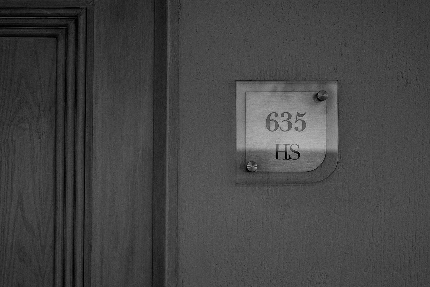 Black and white photo of hotel room number 635 HS sign on wall beside wooden door.