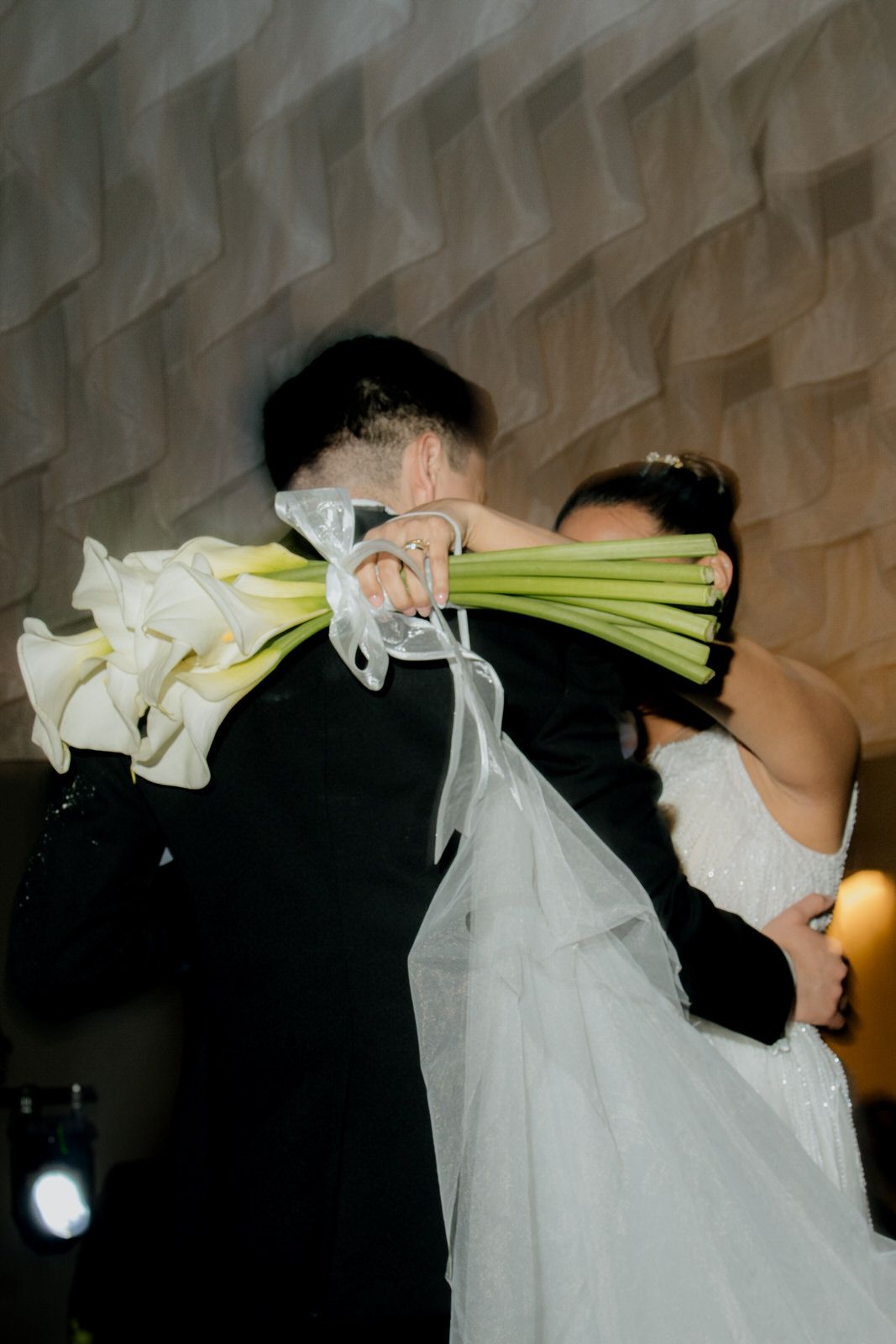 Bride and groom share a romantic embrace, partially hidden behind a bouquet of white calla lilies at their wedding reception.