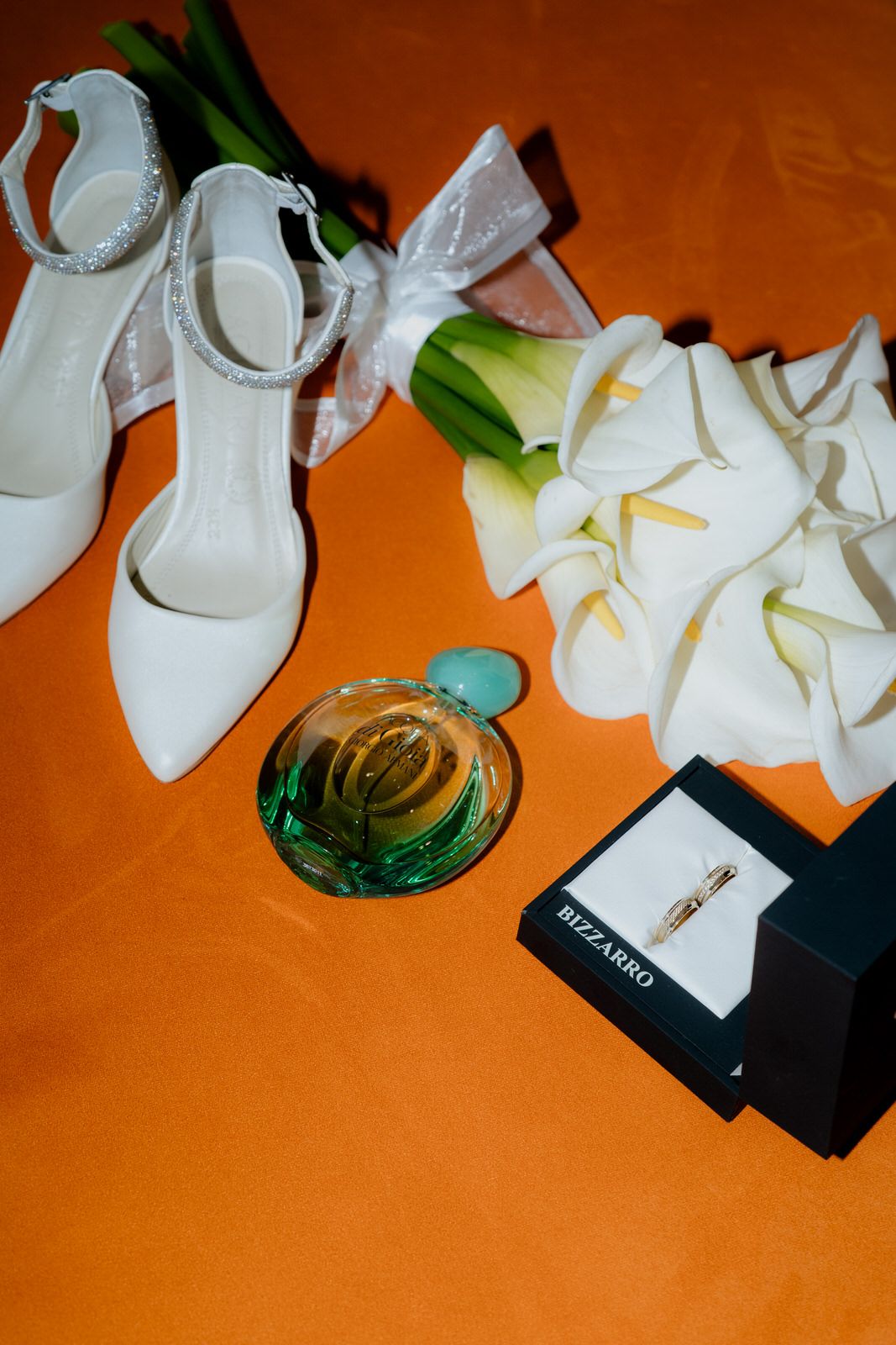 White bridal heels, calla lily bouquet, green perfume bottle, and Bizzarro jewelry box on orange background.