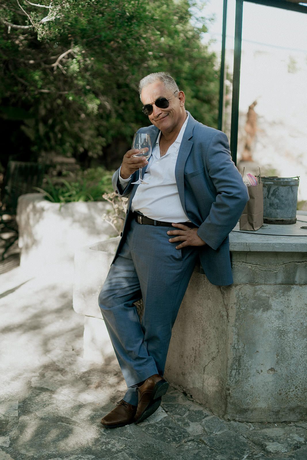 A person in a blue suit and sunglasses relaxes casually on a stone wall in dappled sunlight.