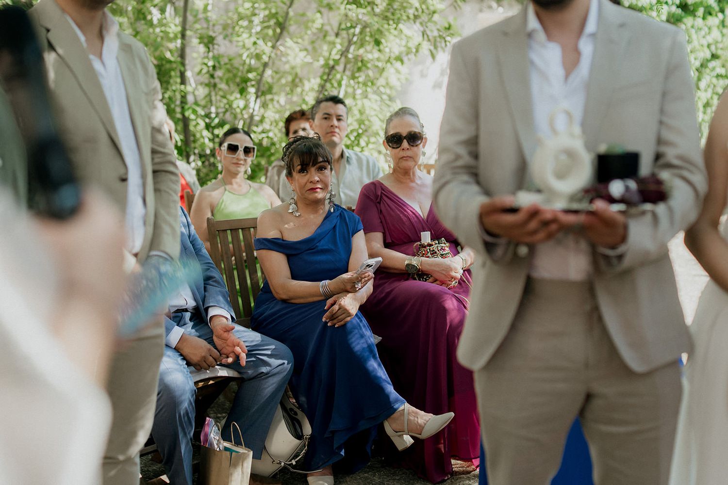 Wedding guests seated outdoors watch ceremony proceedings in a garden setting.