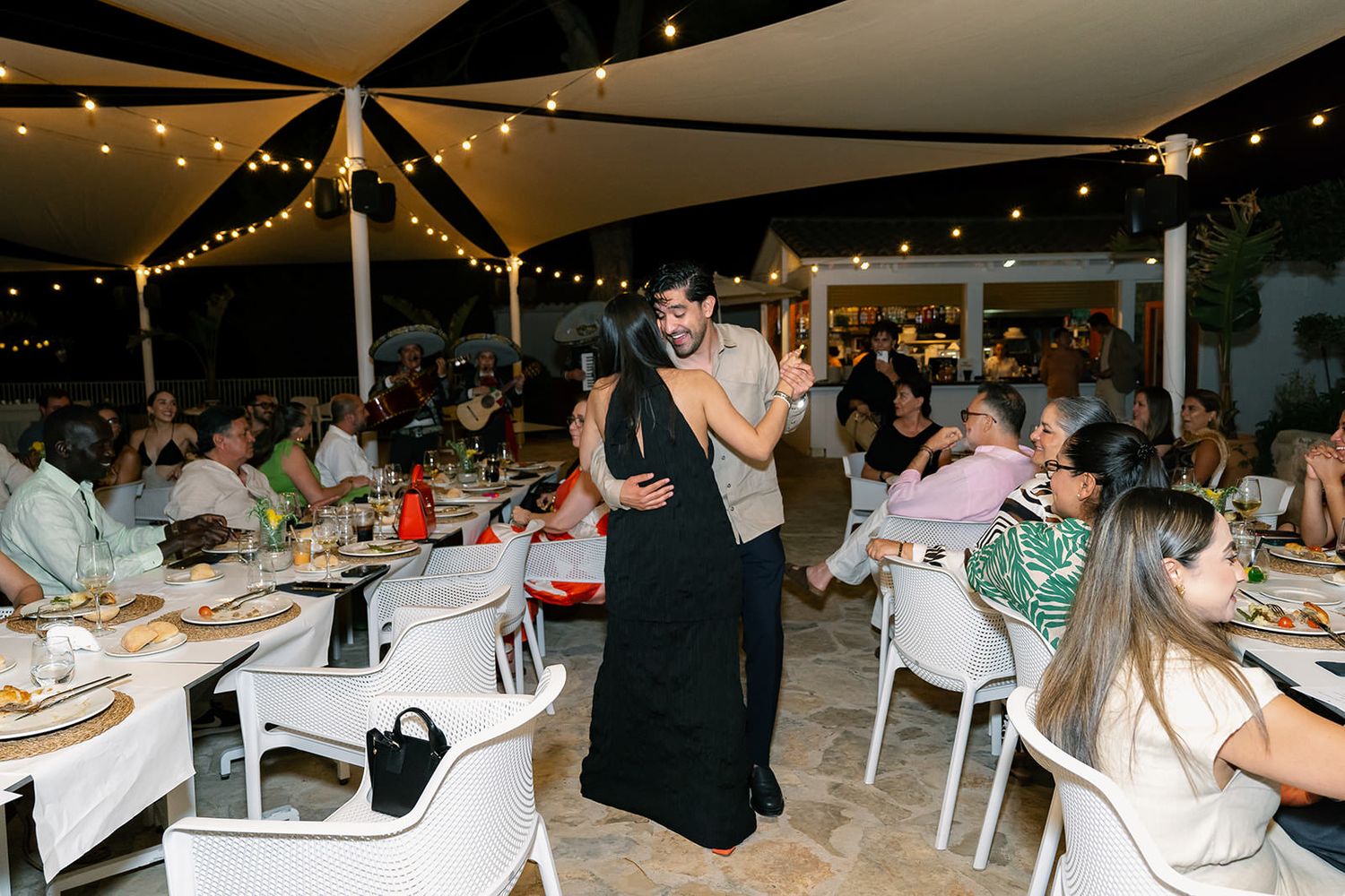 People seated at long tables decorated with string lights for an evening outdoor dinner party.