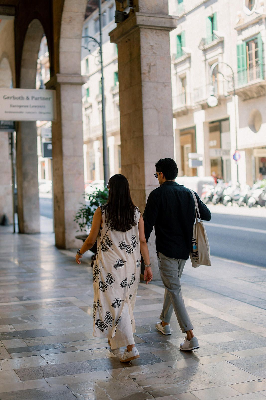 A couple walks hand in hand under stone archways along a European city street.