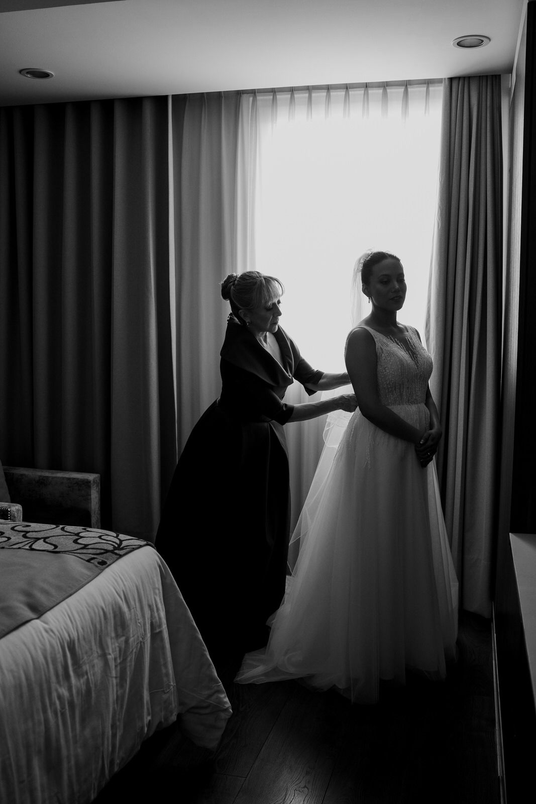 Black and white photo of a bridesmaid helping a bride with her wedding dress in a softly lit hotel room.