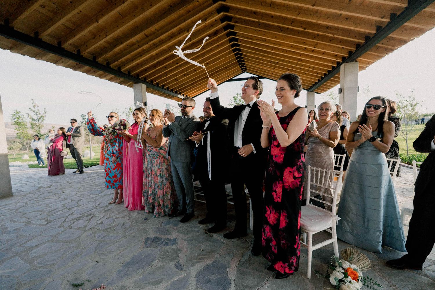 Wedding guests in formal attire stand together under a wooden-beamed pavilion.
