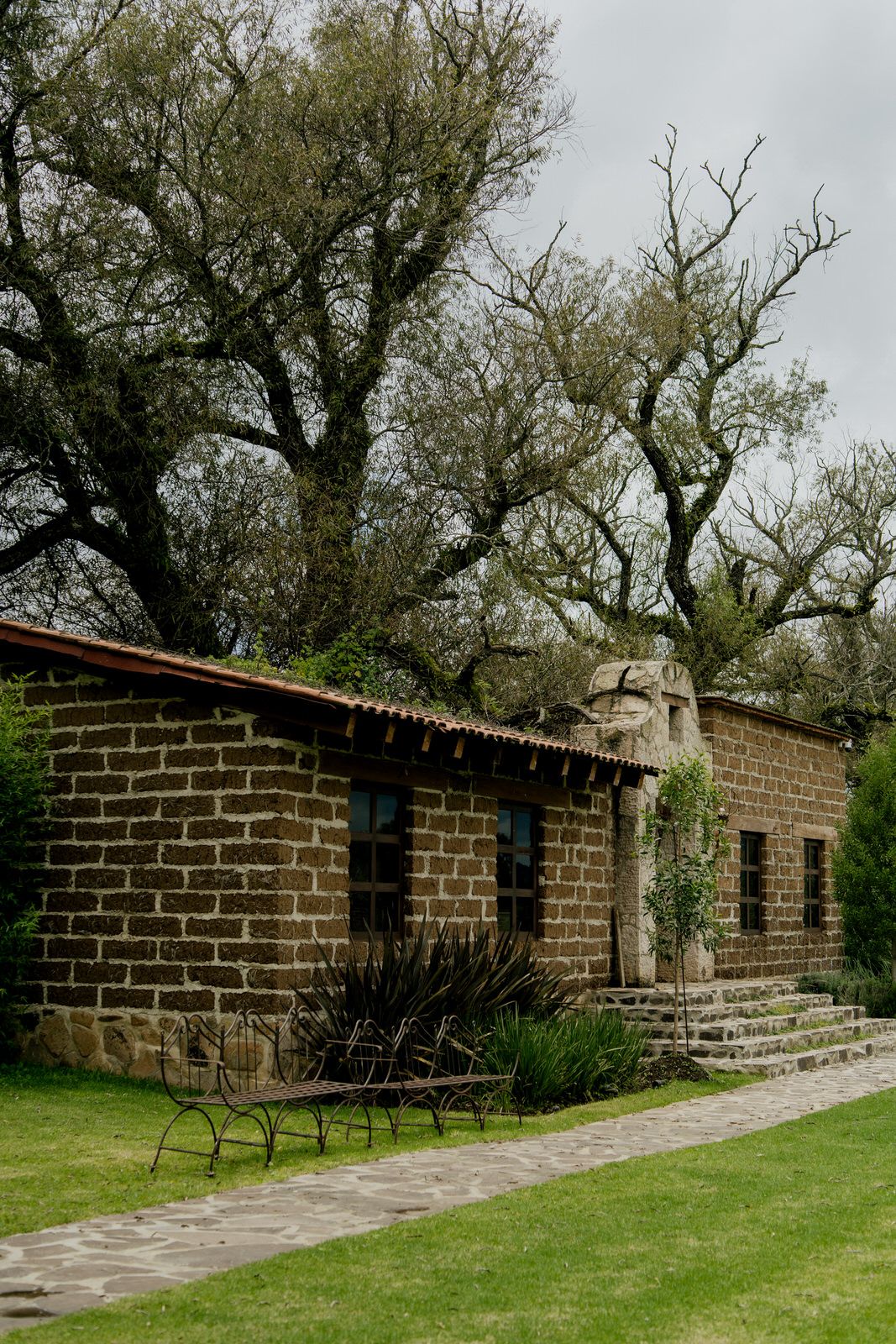 Row of rustic stone buildings with black windows along a paved path on grassy grounds.