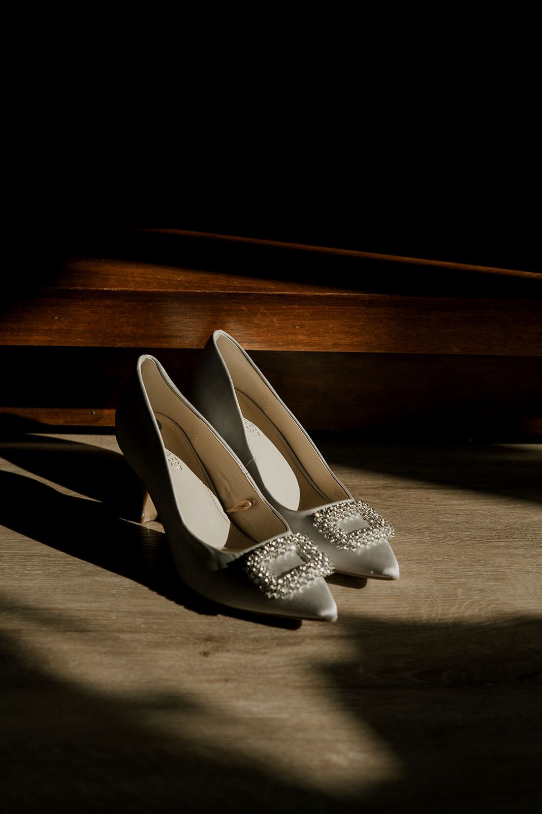 Elegant white bridal heels with pearl buckle detail on wooden floor in dramatic moody lighting.