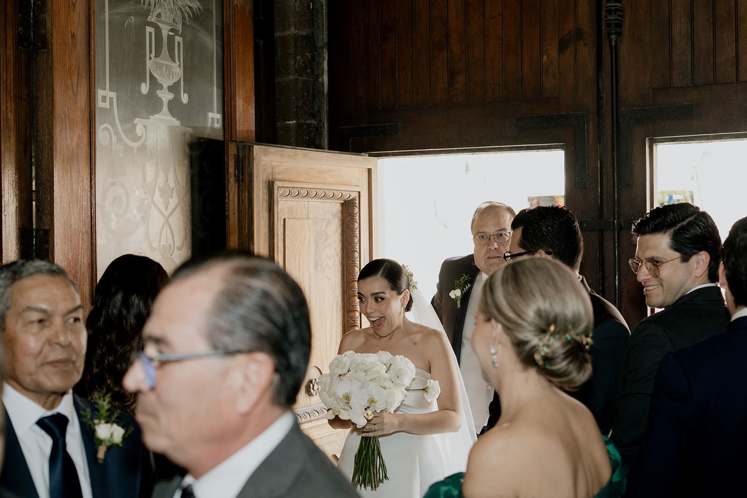 Bride holding white bouquet stands with wedding guests in elegant venue before ceremony.