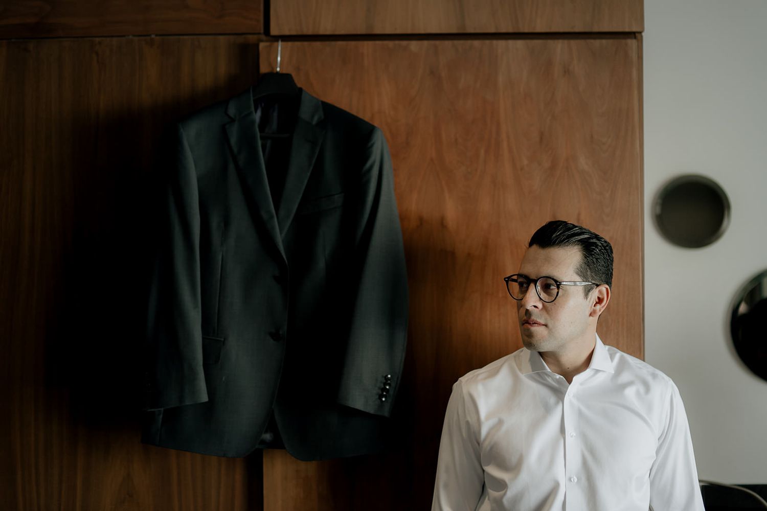 Man in white shirt with glasses stands beside a dark suit jacket hanging on a wooden wardrobe.