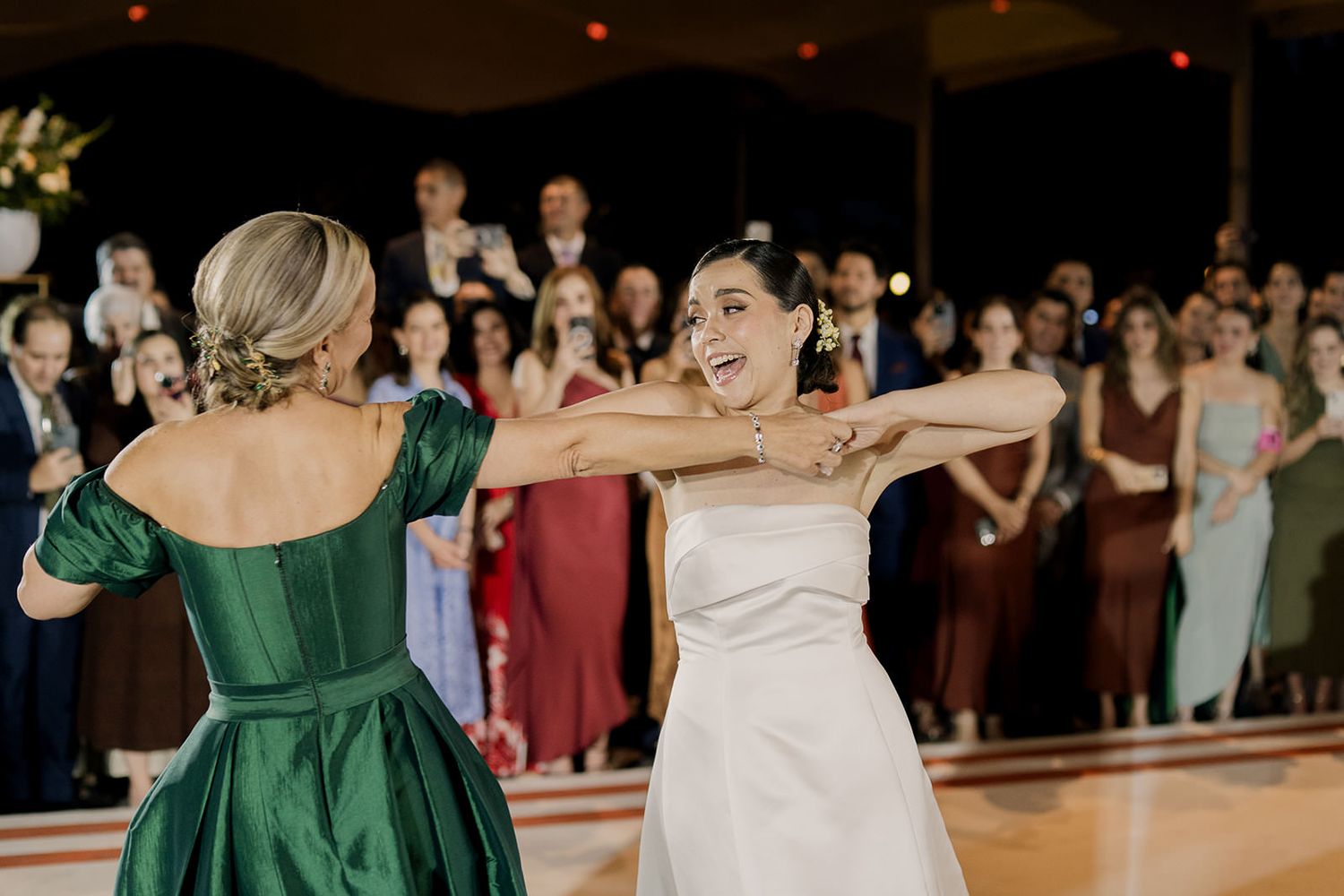 Bride in white wedding gown dancing joyfully with woman in emerald green dress at wedding reception.