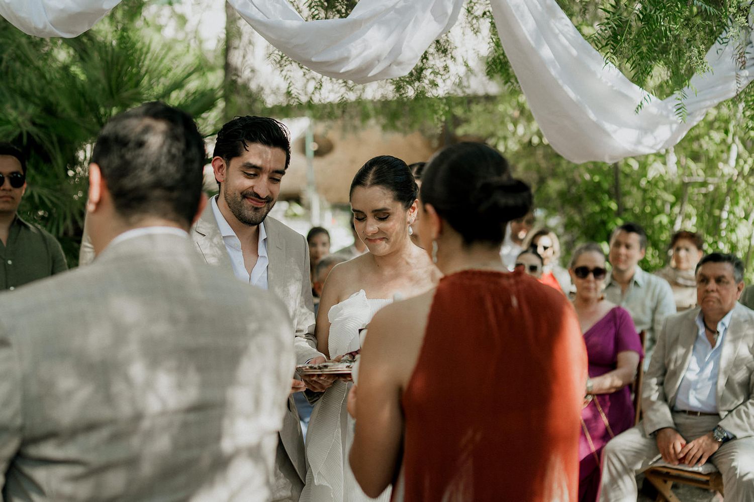 A group of wedding guests gather outdoors under white fabric draping during a daytime celebration.