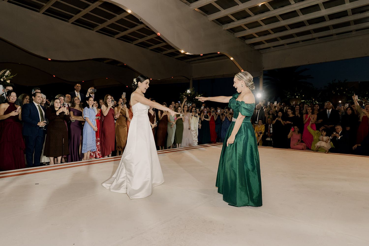 Bride in white gown dancing with woman in green dress on dance floor surrounded by wedding guests.
