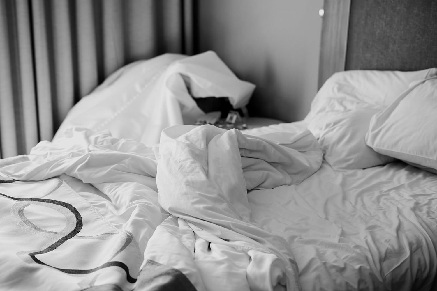 Rumpled white hotel bed with messy pillows and tangled sheets in black and white photography.