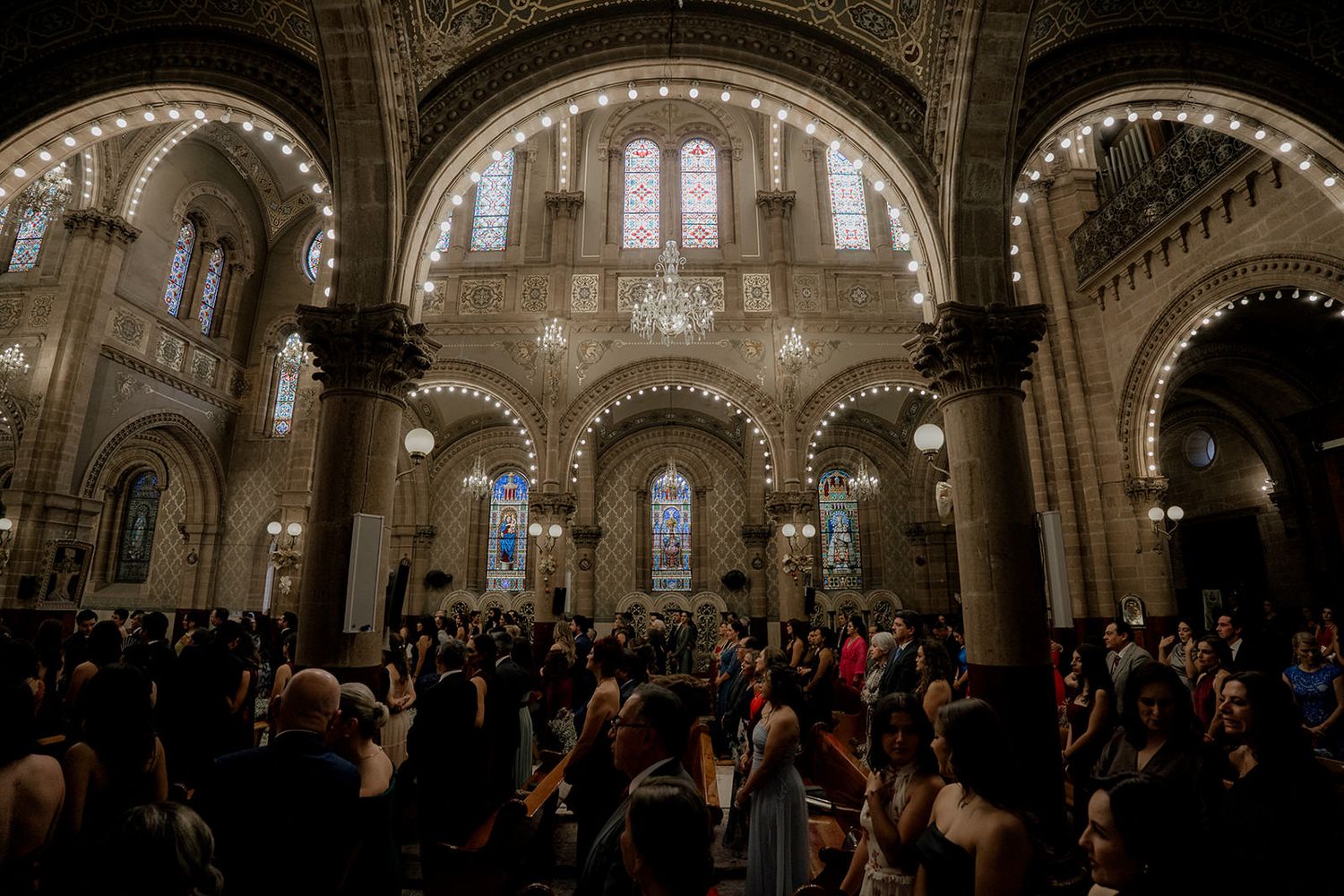 Grand cathedral filled with wedding guests beneath ornate arched ceilings and stained glass windows.