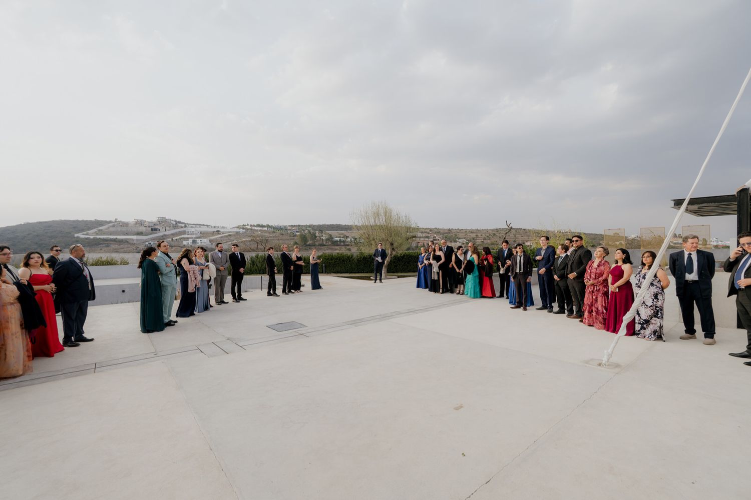 Outdoor wedding ceremony with guests lined up on a rooftop terrace under cloudy skies with scenic hills in the background.
