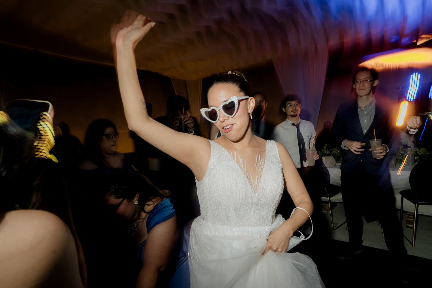 Bride in white gown dancing joyfully wearing white heart-shaped sunglasses at reception.