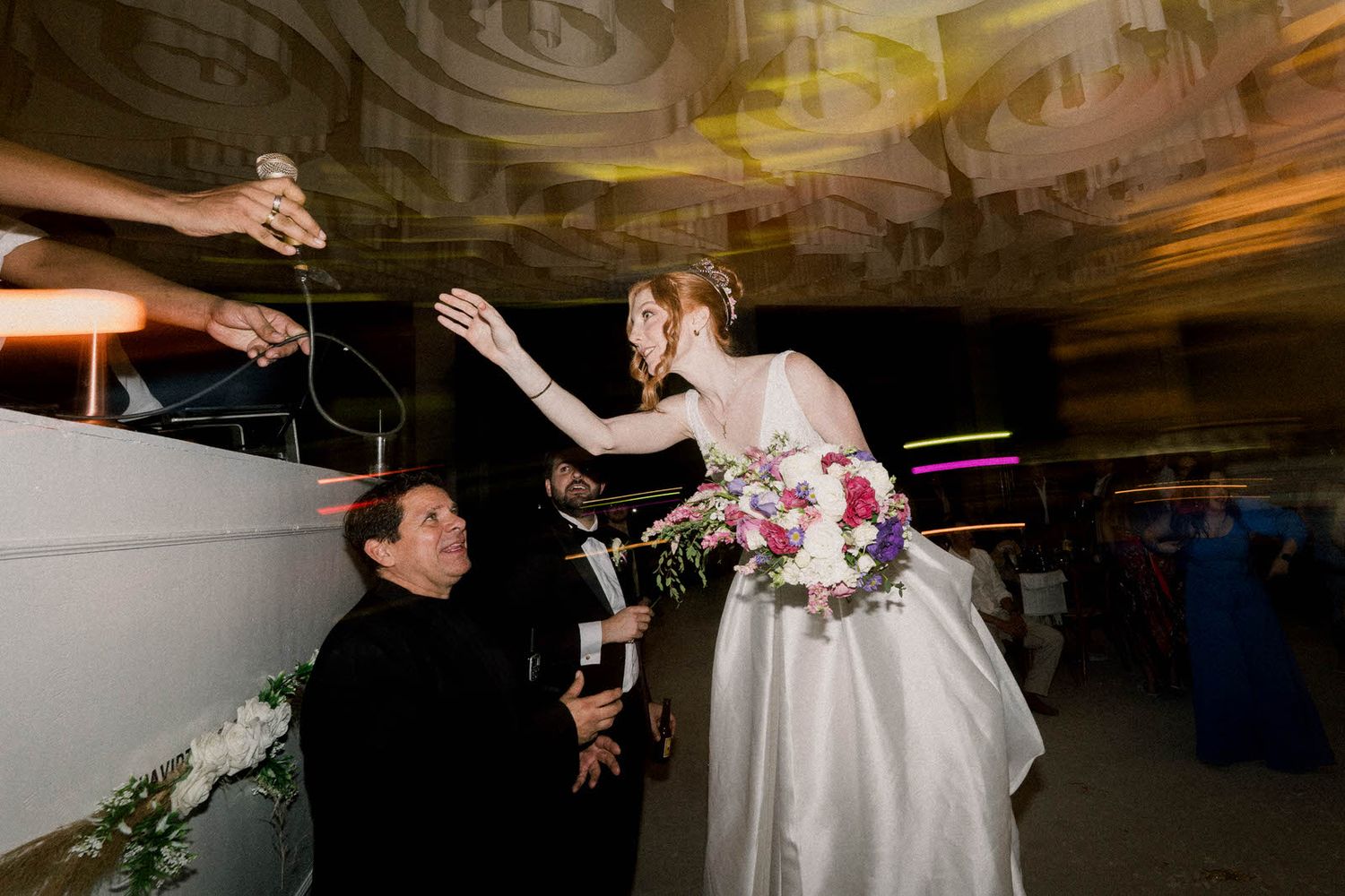A bride in a white gown reaches out to guests while holding a bouquet at an elegant reception venue.