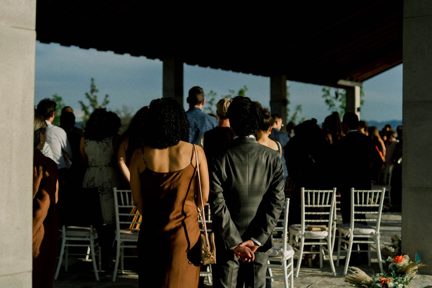 Wedding guests gather under a covered outdoor venue with white chairs at sunset.