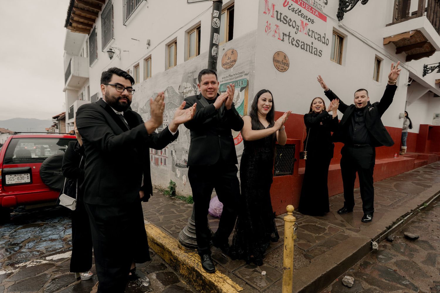Wedding party in black attire celebrates on cobblestone street outside white building.