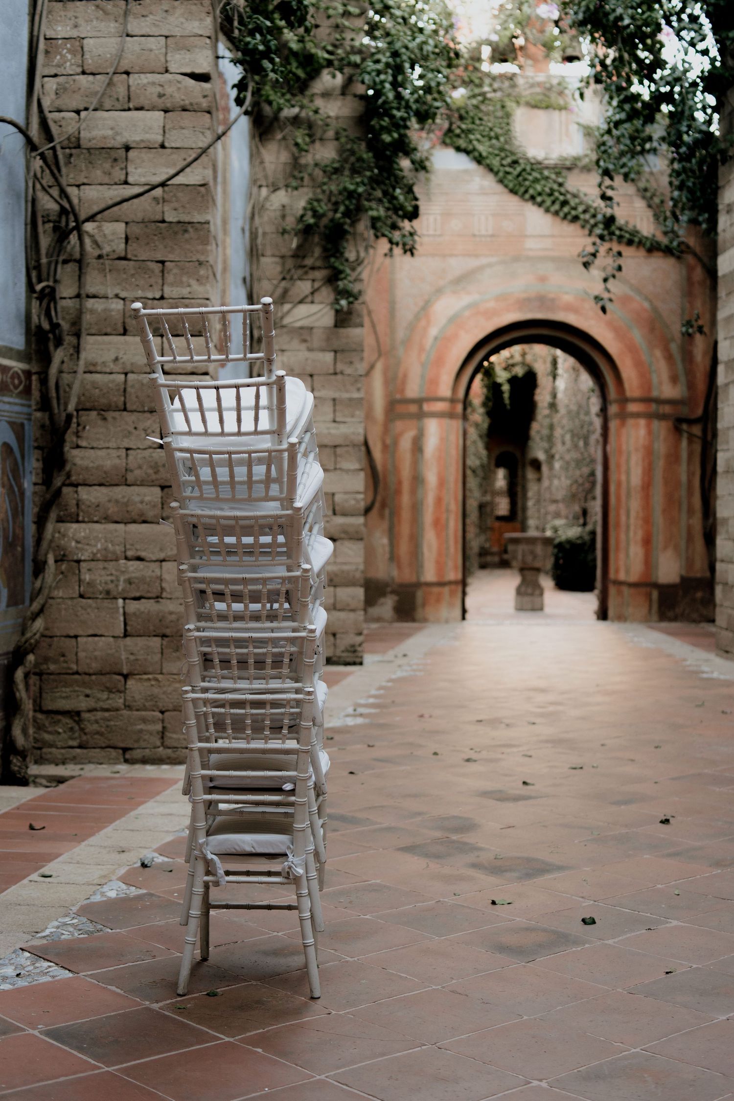 Empty white Chiavari chairs lined up in a stone courtyard with arched doorways.