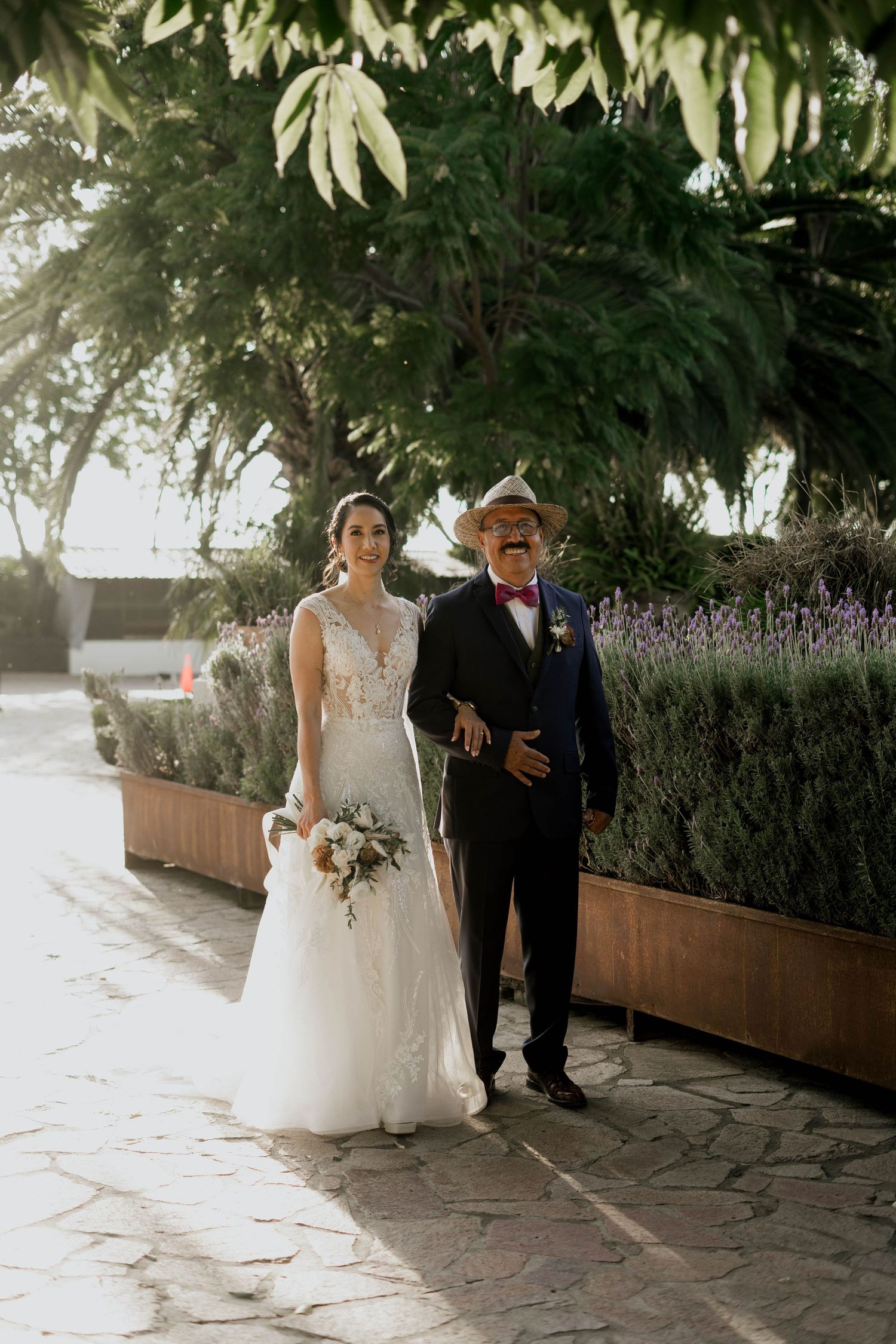 Couple walks along stone pathway under lush trees during wedding ceremony.