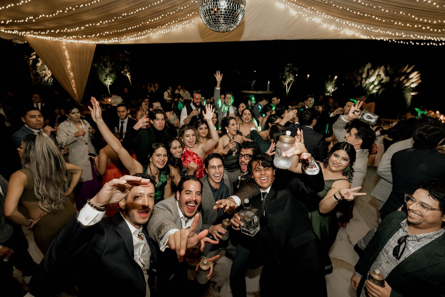 Large crowd of wedding guests dancing and celebrating under disco ball and string lights.