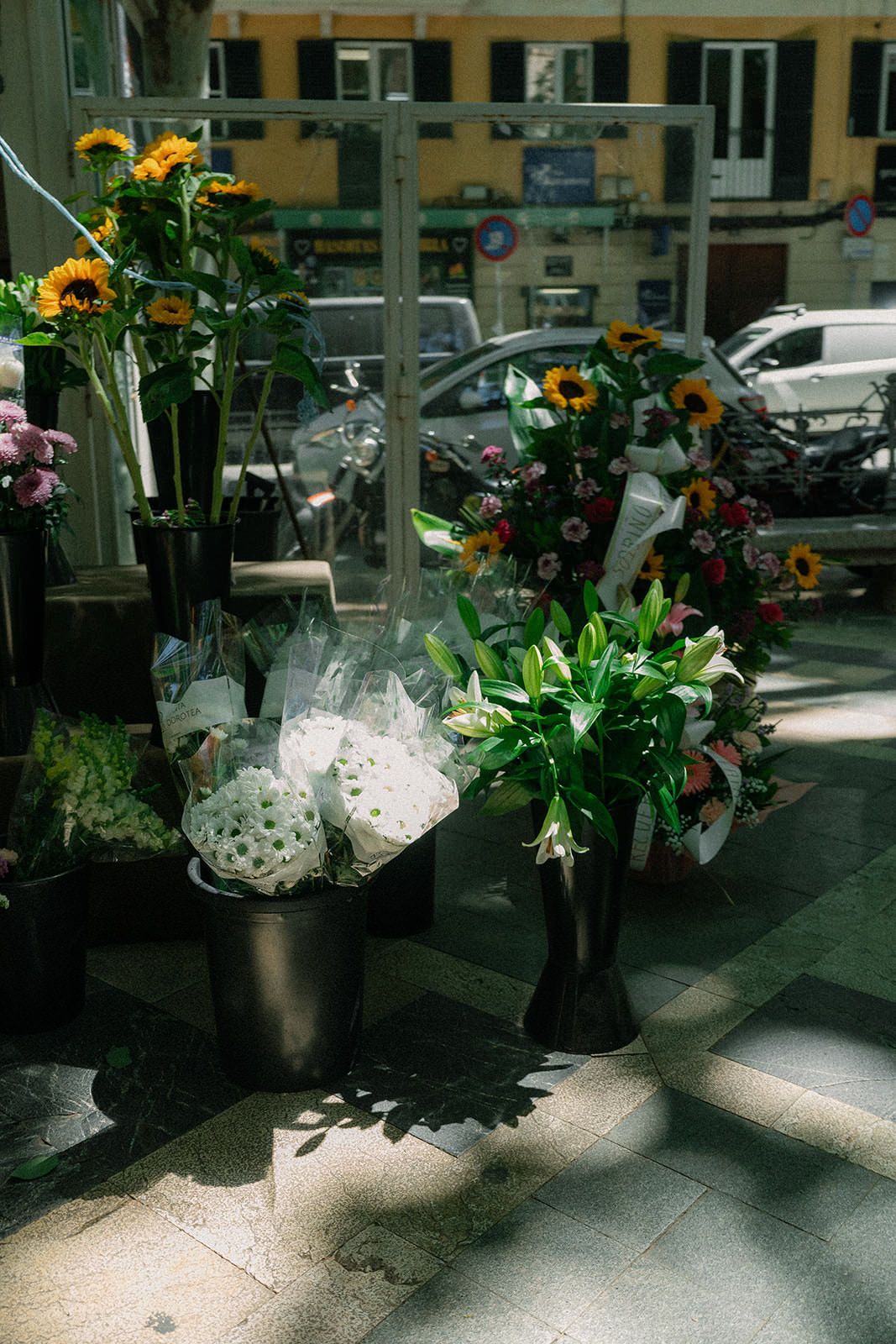Fresh floral arrangements in glass vases displayed on a sunlit floor with cars visible in the background through windows.