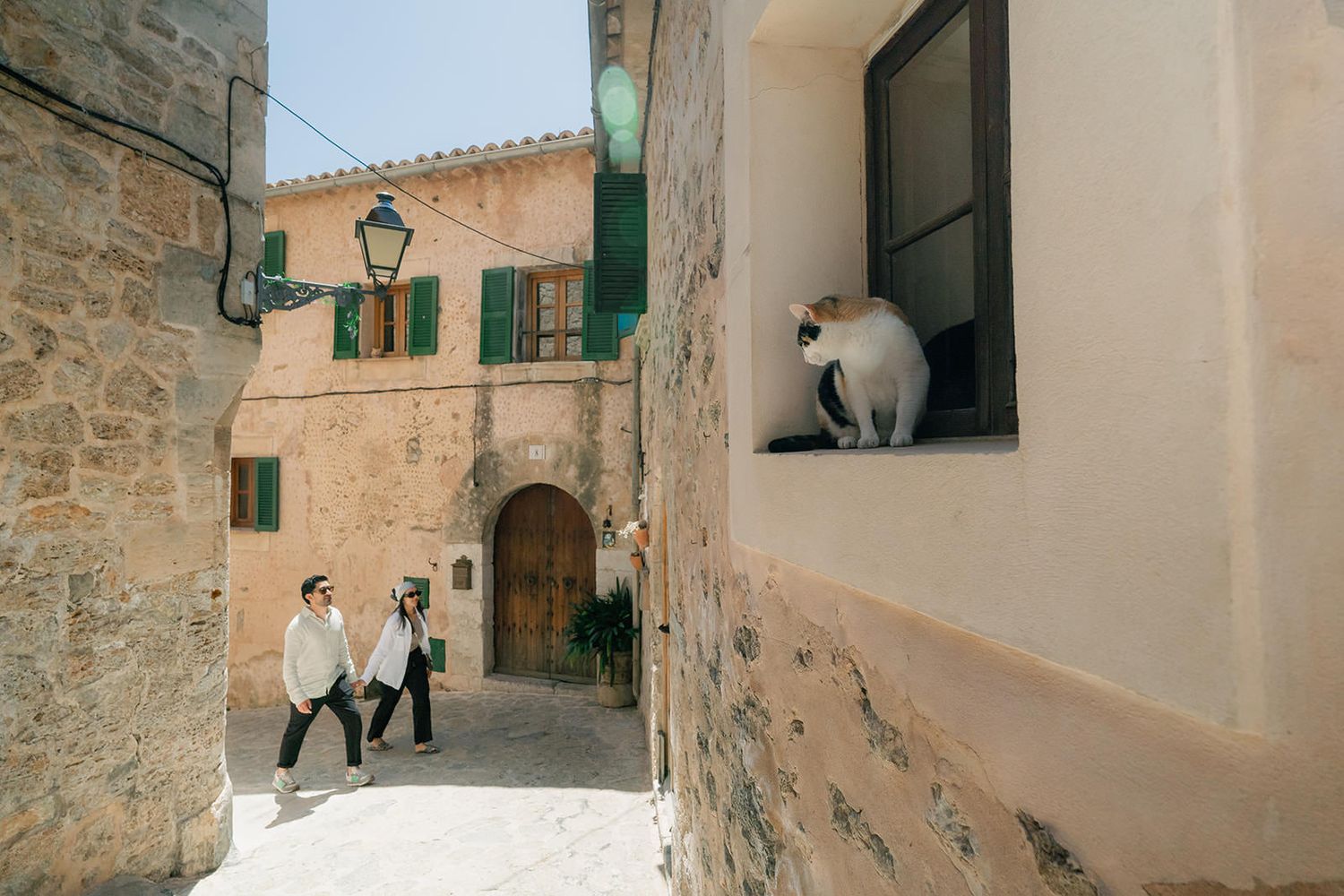 A series of charming alleyways in a Mediterranean village with white walls, green shutters, and a white cat in the window.