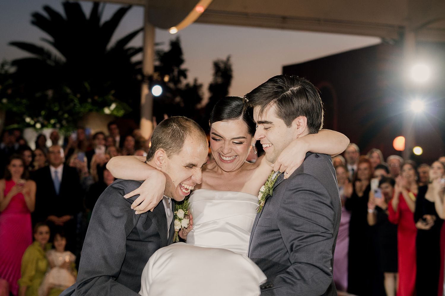 Joyful bride embraced by two groomsmen in gray suits during fun wedding reception celebration outdoors.