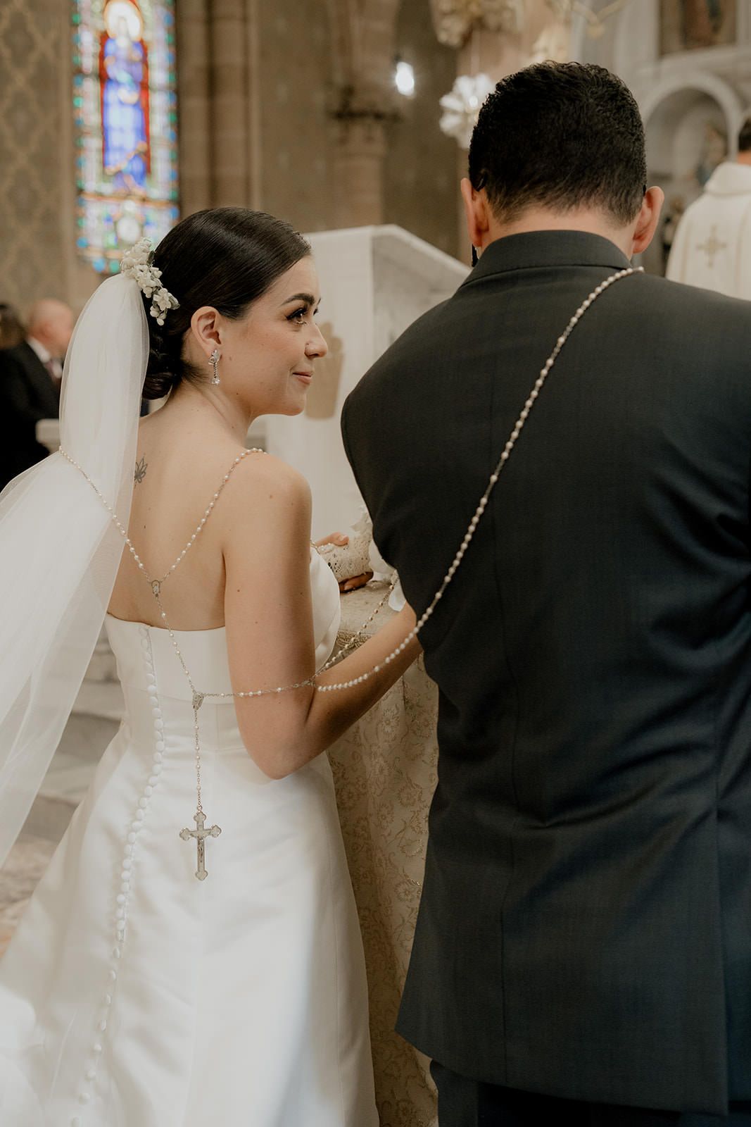 Bride and groom exchanging vows at church altar, bride wearing backless gown with cross pendant and veil.
