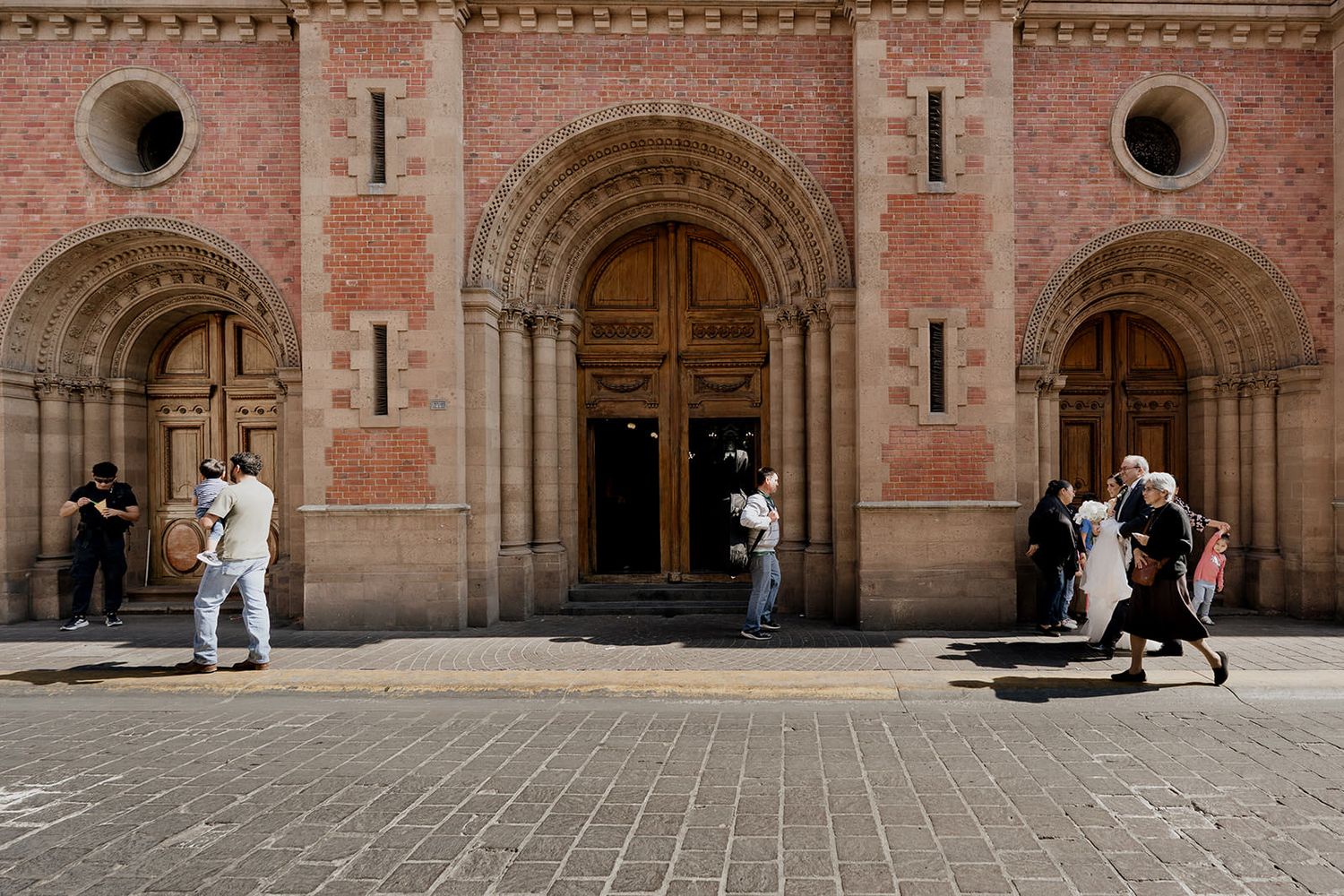 Guests arriving at a historic stone church with ornate Romanesque arched doorways on a sunny wedding day.