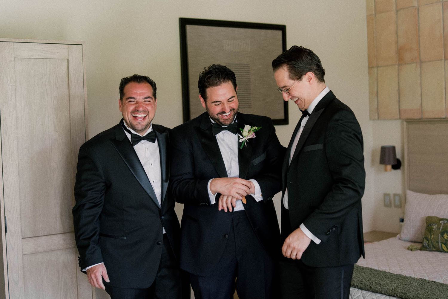 Three groomsmen in black tuxedos share a candid laughing moment while checking a watch before the wedding ceremony.