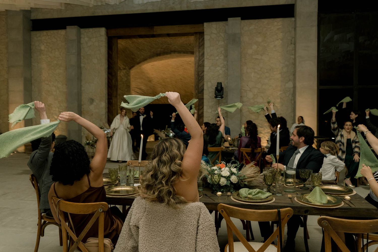 Wedding reception guests celebrate at decorated indoor tables with dramatic lighting and high ceilings.
