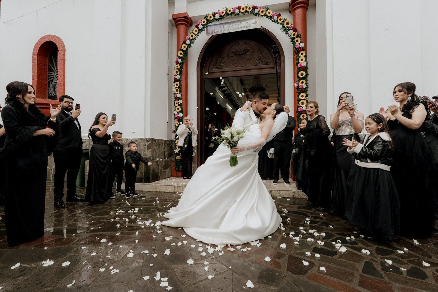 Wedding couple dances outside church entrance while guests in black throw petals in celebration.