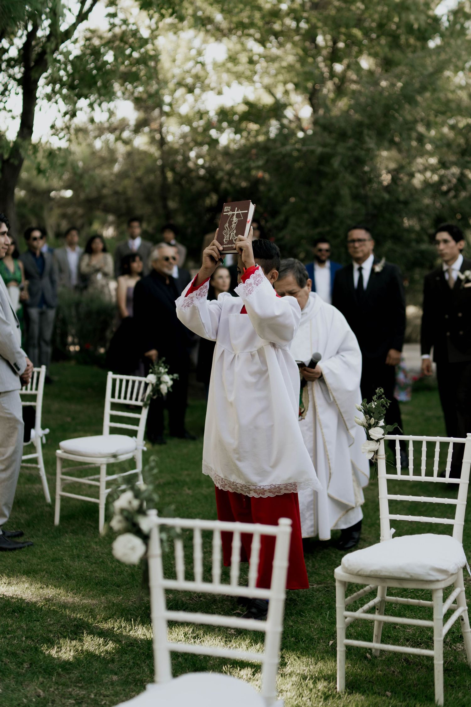 Clergy in white robes raises a ceremonial vessel during outdoor wedding service.