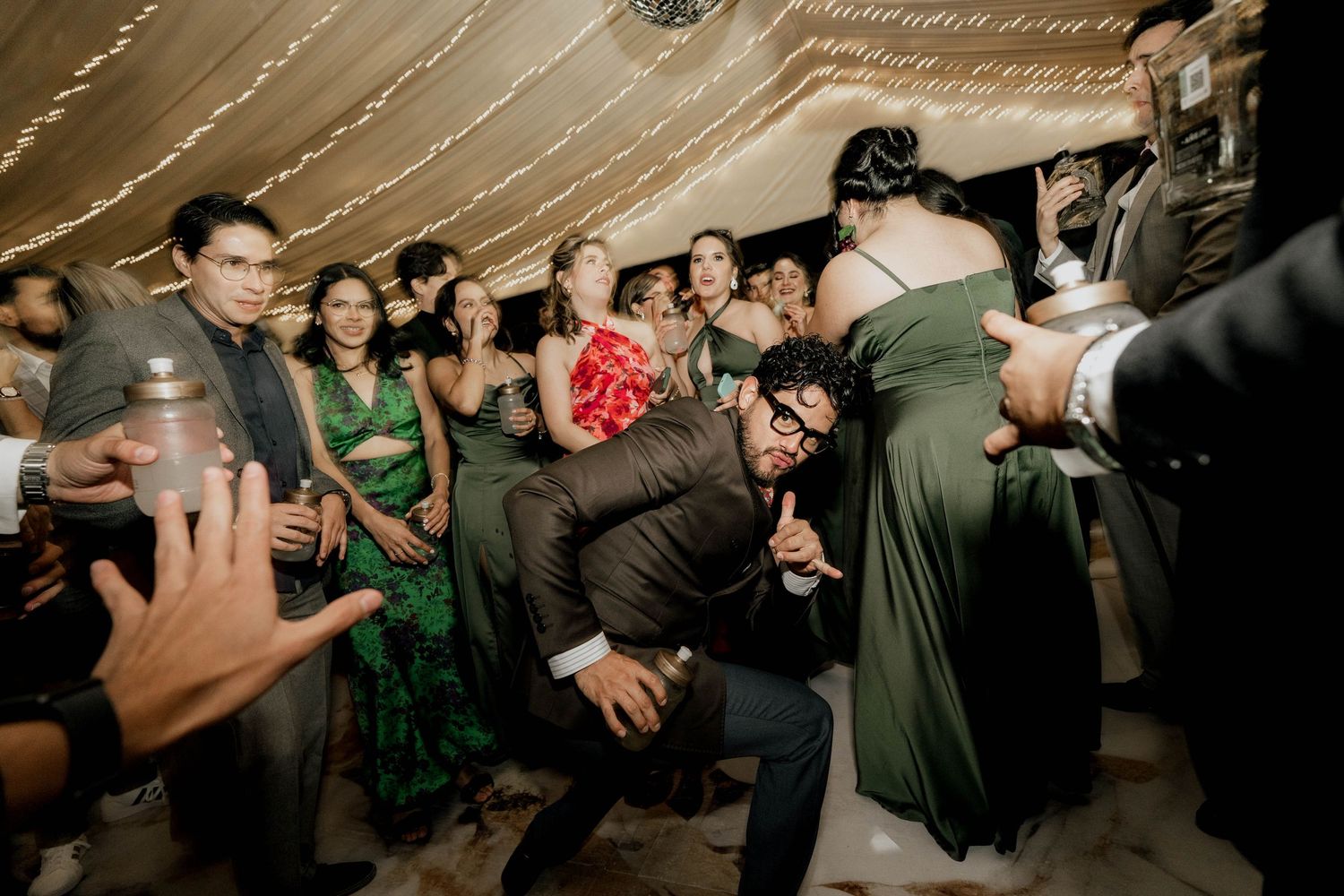 Wedding guests dancing and bending low during lively reception under string lights.
