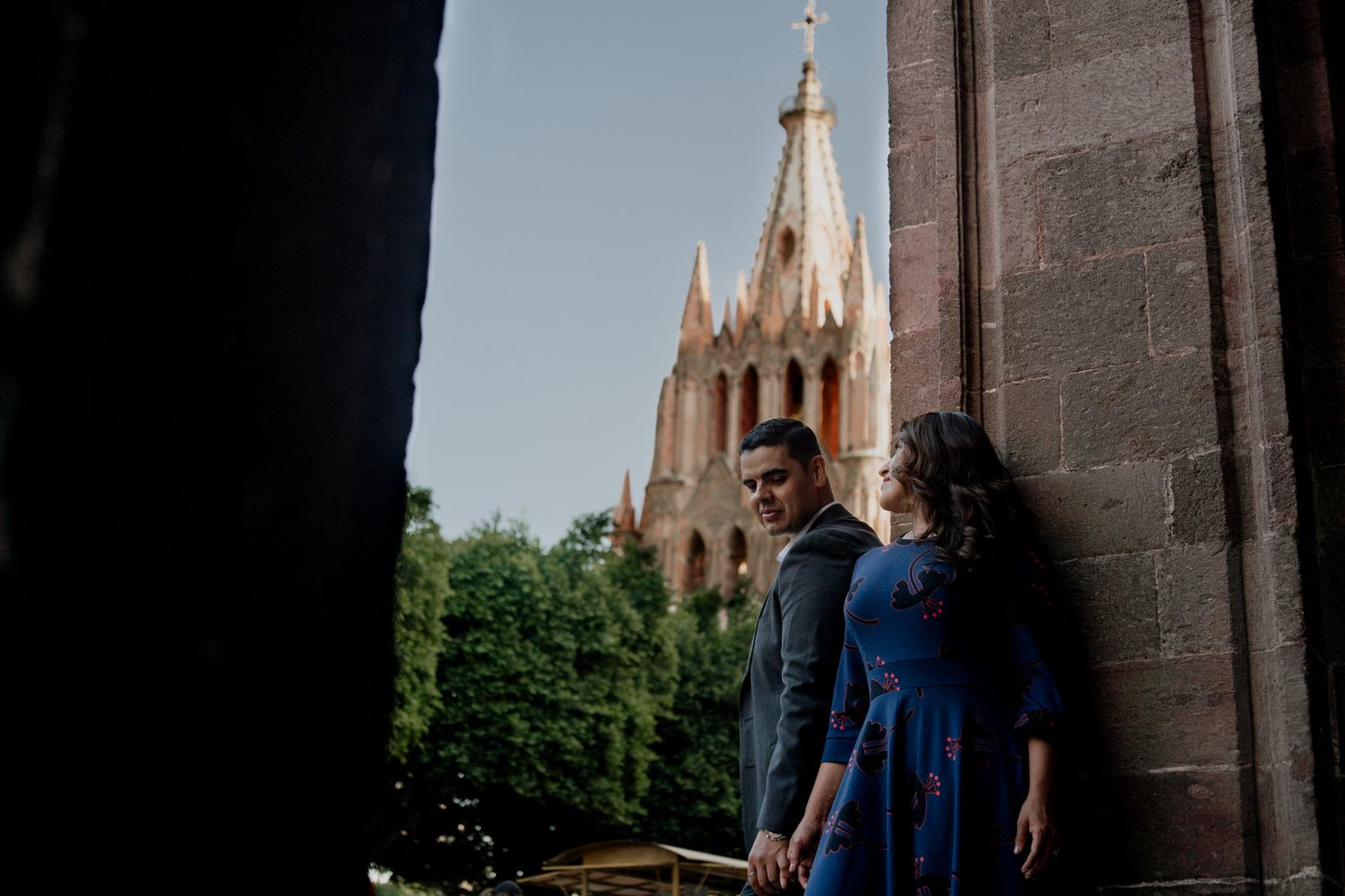 A couple stands in shadow against stone walls with a Gothic church tower in the background.