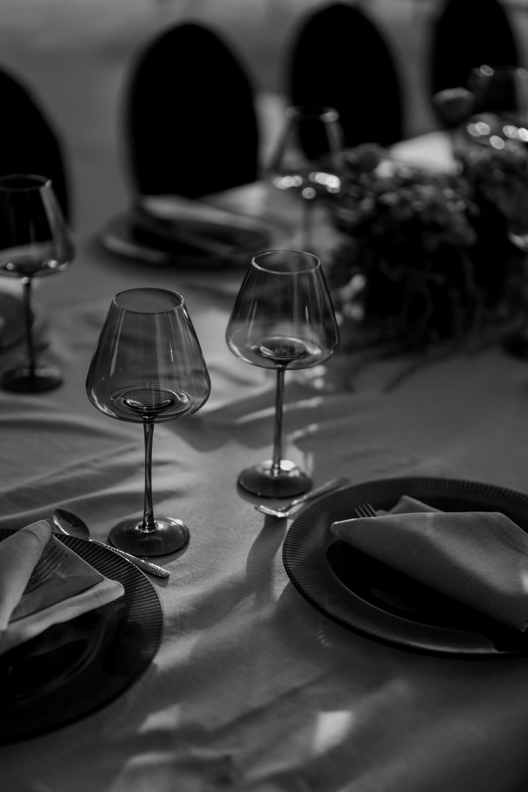 Black and white close-up of a formal table setting with crystal wine glasses, dark plates, and folded napkins with silverware.
